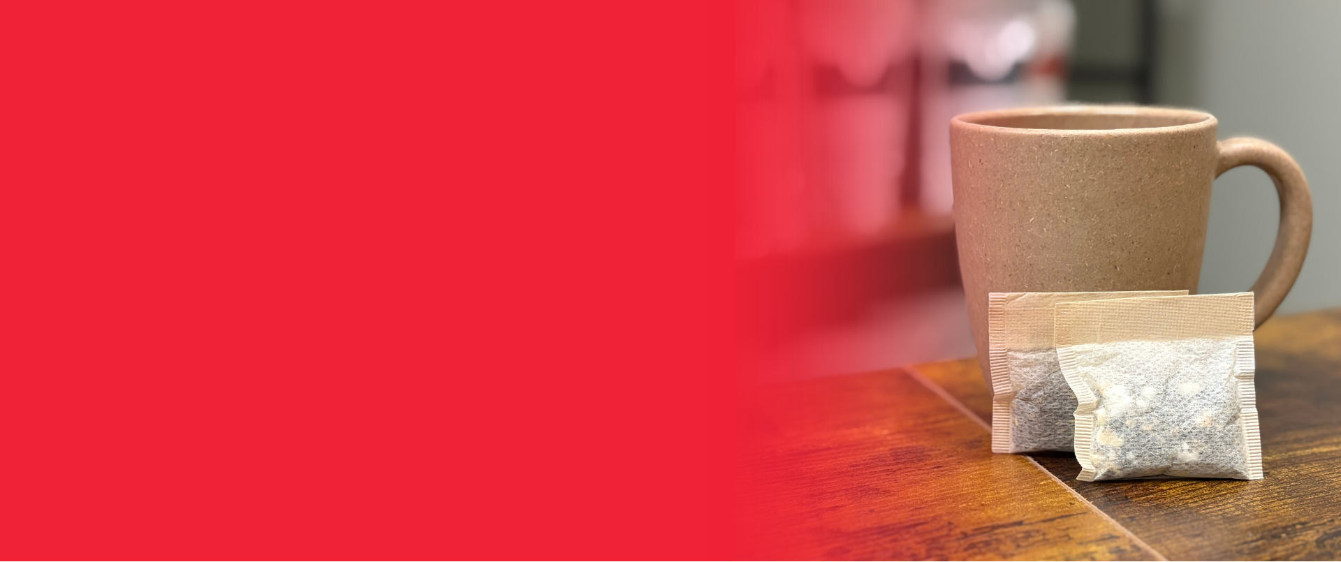 A small paper tea bag packet sits on a wooden table in front of a beige ceramic mug, with a large solid red graphic overlay covering the left side of the frame.